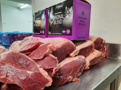 Raw meat on a metal surface with Orgreenic Pets Original Blend cat food packaging in the background.